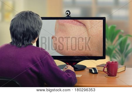 Telemedicine female dermatologist examines mole in the right side of womans chin on computer monitor carefully. Virtual doctor looks at nevus on the face either by online video chat or snapshot. Horizontal mid-shot on blurry indoors background