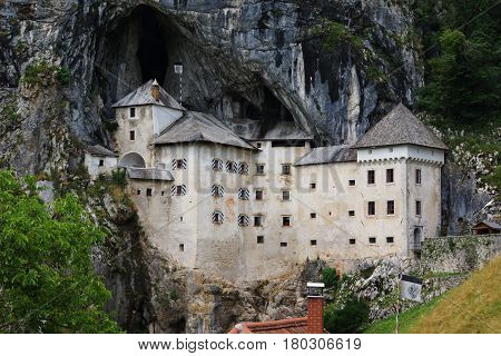 fifteenth-century Predjamski castle built into the cave in a rocks with a height of over one hundred meters. White with sloping roofs on a background of black rocks and green trees.