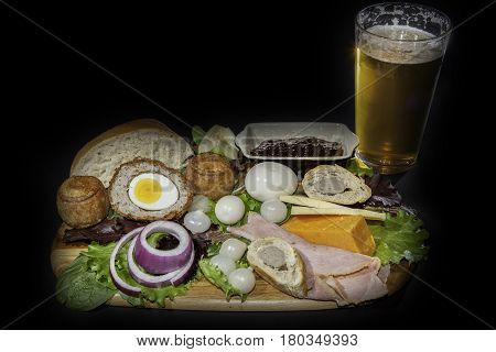 Ploughman's pub lunch including scotch egg; pork pies ham; pickled onions and cheese served with a pint of lager beer. Isolated against a black background.