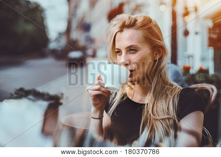 Beautiful adult blonde woman drinking enjoying delicious hot tea and thoughtfully looking outside while sitting in street cafe alone on sunny summer evening after hard work day