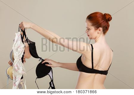 Bosom concept. Slim attractive red hair woman wearing black underwear holding many bras in hand choosing witch bra to wear