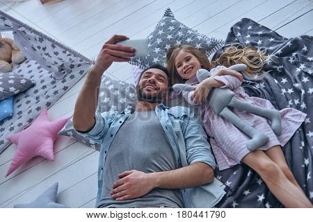 Capturing bright moments together. Top view of cheerful father and his little daughter taking selfie while lying on the floor at home together