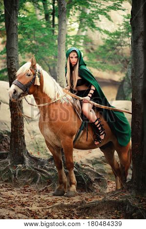 Vertical shot of a sexy brave female elf archer in a green cape riding her horse in the woods danger dangerous skilled fighter weapon armed mysterious forest hiding hunting costume character creature.