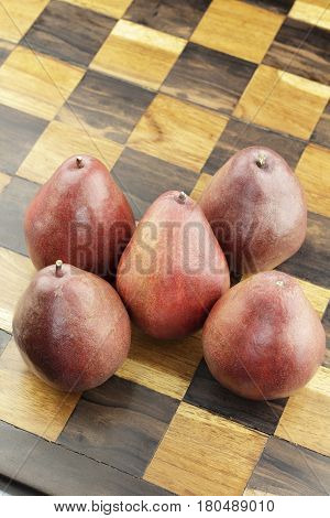 Five organic red pear fruit on inlaid wooden chessboard close-up. Handmade inlay brown and beige wood chess set board with several organic red pears on its surface.