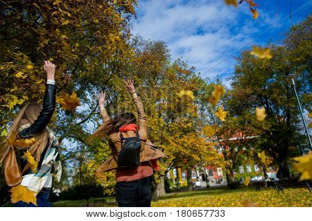 Mellow autumn. Two girls are students cheerfully spend time in the city park. Good weather blue sky. Girlfriends throw armfuls of dry yellow leaves up and rejoice as children.