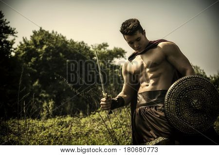 Young handsome muscular man posing in roman or spartan gladiator costume with shield and sword on field.