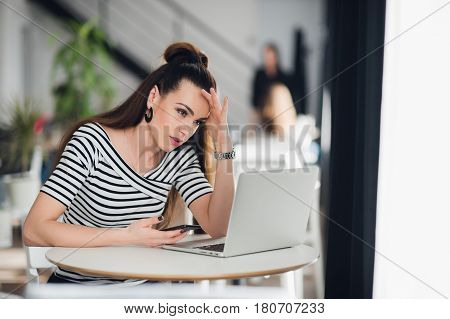 Worried and sad woman got bad news from her relatives in a cafe. Puzzled female looking at a laptop and holding phone