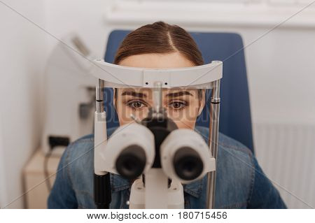 Test for myopia. Pleasant attractive young woman sitting in the chair looking into binoculars while having an eye test for myopia