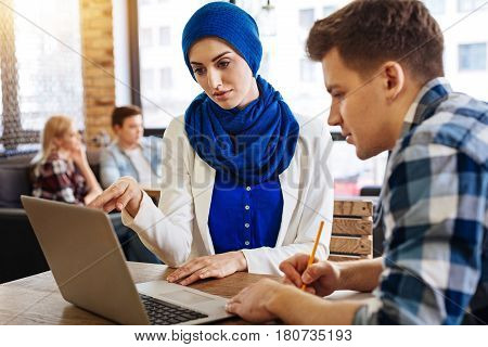International program. PLeasant nice muslim woman sitting at the table with her groupmate and using laptop while getting ready for classes