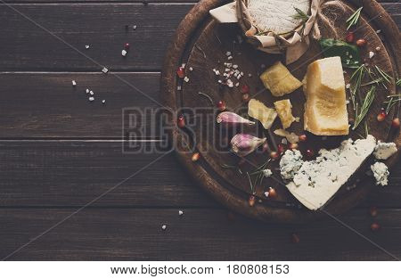Cheese delikatessen top view on rustic wood. Wooden round desk with blue cheese roquefort, parmesan, camembert and brie cuts decorated with garlic, pomegranate and basil, french cuisine ingredients