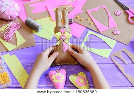 Small child holds a felt Easter bunny in his hands. Child made a felt cute bunny with hearts for Easter. Tools and materials for art creativity on a wooden table. Simple spring crafts project for kids