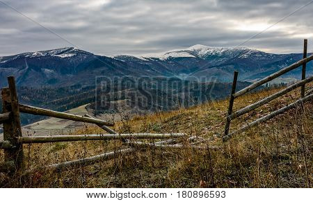 wooden fence on the hill with weathered grass. mountains with snowy peaks in the distance. Late autumn landscape in cloudy weather.