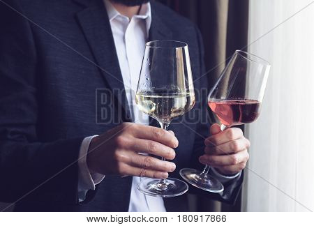 Horizontal close up of Caucasian man in black suit and white shirt offering two tall glasses with rose and white wine at an event by the window natural lighting