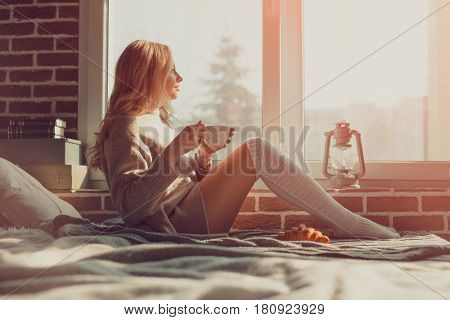 Side view of a woman having a hot drink and cornetto sitting on the bed.