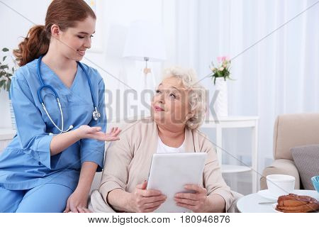 Nurse helping elderly woman to work on tablet pc in light room