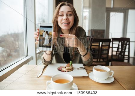 Smiling woman showing photo to her friend using phone while they eating in cafe