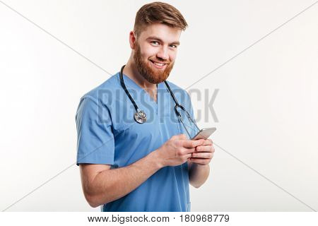 Portrait of male doctor texting, loooking at camera and smiling while standing against white background.