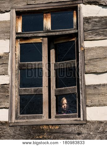 Small and lonely caucasian baby girl or toddler looking out of old wooden cabin window to suggest poverty or recession
