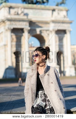 Attractive young woman wearing gray coat with long dark hair and sun glasses posing outdoor in Milan streets, Italy. Beautiful caucasian model portrait. Steet fashion