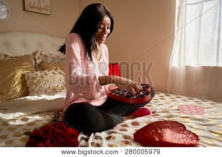 happy african american woman eating romantic valentines chocolates from heart shaped box while sitting on bed at home