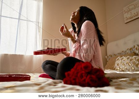 happy african american woman eating romantic valentines chocolates from heart shaped box while sitting on bed at home