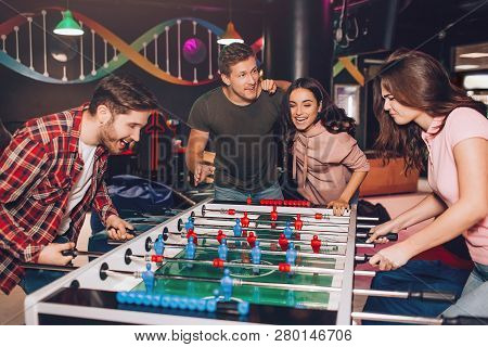 Enthusiastic Excited Young Man And Woman Playing Table Soccer In Room. Their Friends Stand Beside An