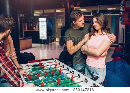 Upset Young Woman Stand At Tabel Soccer In Playing Room. Guy Try To Comfort Her And Embrace. They St