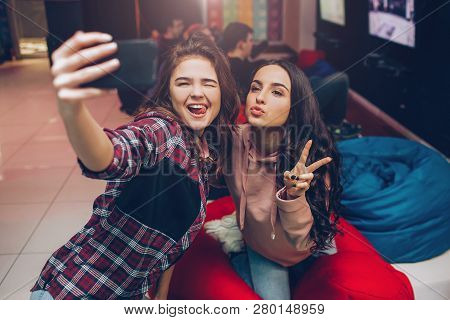 Two Young Women Taking Selfie On Phone Camera And Posing In Playing Room. They Shink And Show Finger