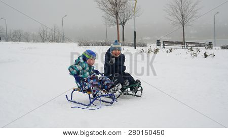 Two Little Children Sledding On The Snow-covered Road In Winter Park In The Sunless Afternoon. Snow 