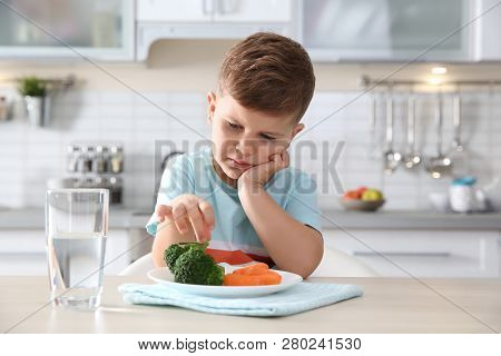 Unhappy Little Boy Eating Vegetables At Table In Kitchen