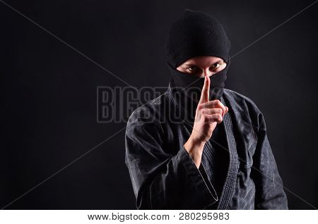 Quiet! Shh Symbol. Criminal Ninja In Balaclava On A Dark Background Holds Index Finger On Lips.