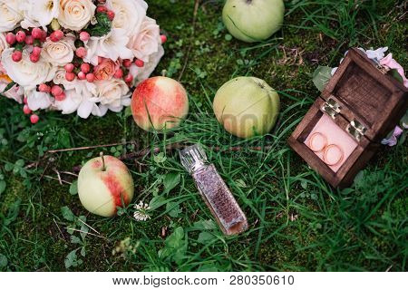 Wedding Rings In A Wooden Box In The Grass With Apples And Perfumes With A Wedding Bouquet Of Flower