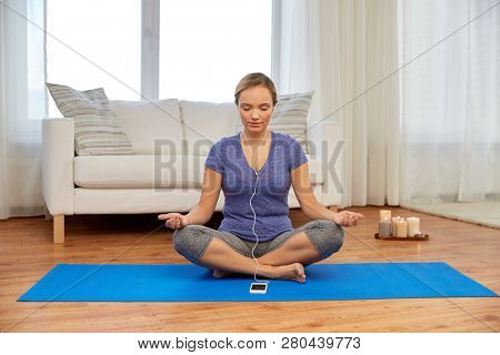 yoga, technology and mindfulness concept - woman with smartphone and earphones meditating in lotus pose at home
