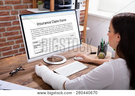 Businesswoman With A Plaster Cast Hand Filling Insurance Claim Form On Computer In The Office