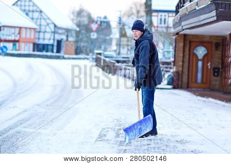 Man With Snow Shovel Cleans Sidewalks In Winter During Snowfall. Winter Time In Europe. Young Man In