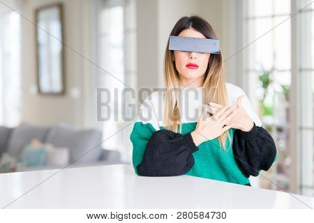 Young beautiful woman blindfold eyes with black censor band at home smiling with hands on chest with closed eyes and grateful gesture on face. Health concept.