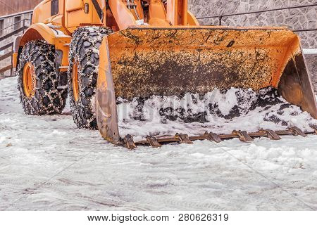 An Orange Grader With Snow Chains At The Wheels And A Big Bucket On A Road In Winter