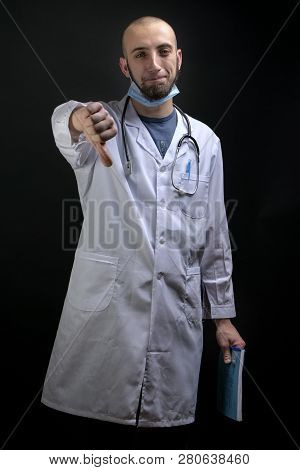 Young Male Doctor Holding A File, Having Stethoscope And Mask Displaing A Finger Thumb Down. Bearded