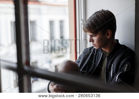 Cute Handsome Young Guy With A Fashionable Hairstyle In Stylish Clothes Sitting On The Window Indoor