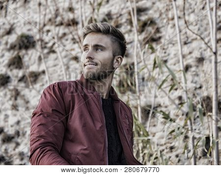 Handsome Young Man Outdoor In Winter Fashion, Wearing Red Jacket And Woolen Scarf In City Park
