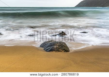 A Rock On A Sandy Beack With Watter Foaming Around It And Waves Coming In From The Ocean