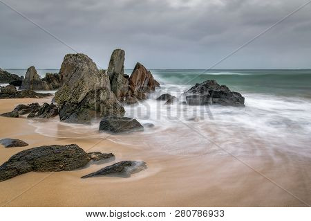 Blurred Waves Swirling Around Rocks On A Sandy Beach In Ireland.  The Sky Is Stormy But The Blurred 