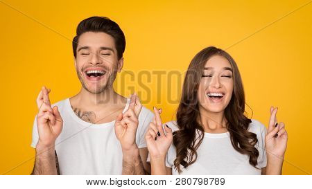 Cheerful Couple Cross Fingers With Hope For Good Luck On Yellow Stusio Background, Crop