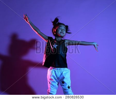 Joyfull Asian Kid Girl In Shirt And Pants With Stars And White Sneakers On Purple Background Dances 