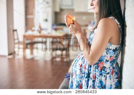 Side View Of Pregnant Woman In Dress Standing In Kitchen And Keeping In Hands Cigarette And Lighter.