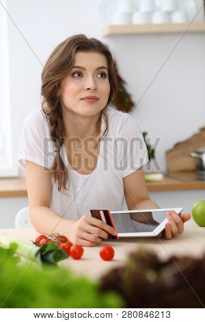 Young  Woman Looking For A New Recipe For  Cooking In A Kitchen. Housewife Is Making Online Shopping