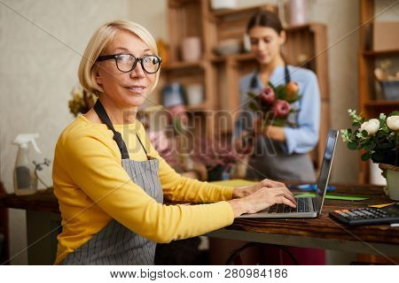 Side View Portrait Of Mature Female Small Business Owner Using Laptop And Looking At Camera In Flowe
