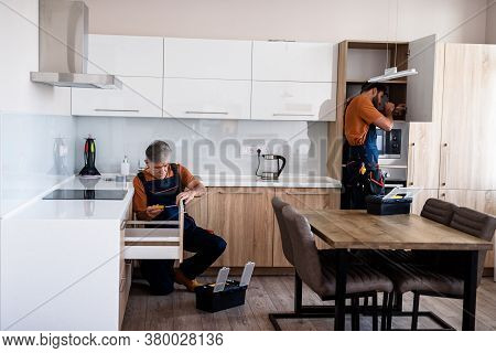 Full Length Shot Of Two Handymen, Workers In Uniform Assembling Kitchen Cupboard, Cabinet Using Scre