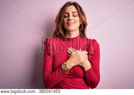 Young beautiful brunette woman wearing casual sweater stnding over pink background smiling with hands on chest with closed eyes and grateful gesture on face. Health concept.