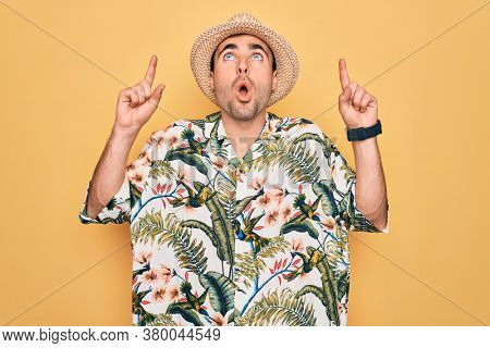 Young handsome man with blue eyes on vacation wearing summer florar shirt and hat amazed and surprised looking up and pointing with fingers and raised arms.